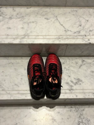 Nike Air Max Plus röd/svart - Nike Air Max Plus sneakers i rött och svart med klassisk Tn-detalj på hälen. Skorna har svarta snören, mesh- och syntetmaterial samt den ikoniska vågiga designen på ovandelen. Orange swoosh på sidan ger extra attityd. Perfekta för dig som gillar streetwear.