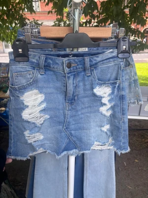 Blå jeanskjol med slitningar - Säljer en kort blå jeanskjol från Hollister med slitna detaljer och rå kant nertill. Kjolen har klassisk femficksdesign, bälteshällor och knappstängning framtill. Perfekt för dig som gillar en avslappnad och trendig stil.