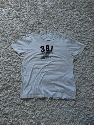 381 | Polo Ralph lauren - Vit t-shirt med tryck i vinrött och grått framtill, där det står 381 och PROPERTY 258. Klassisk rund hals och korta ärmar. Perfekt till jeans eller shorts för en avslappnad look. Tröjan är från polo Ralph lauren och i princip helt oanvänd.