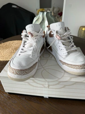 Vita Air Jordan 3 med rosa detaljer - Snygga Air Jordan 3 sneakers i vitt läder med ljusrosa och bruna detaljer. Skorna har klassisk siluett, mönstrad mudguard och rosa snörlås. Perfekta för dig som vill sticka ut med stilrena och trendiga sneakers.