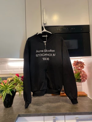 Svart zip hoodie Acne Studios - Säljer en svart hoodie med dragkedja från Acne Studios. På bröstet står det 'Acne Studios STOCKHOLM 1996' i vitt tryck. Hoodien har långa ärmar, ribbade muddar och är tillverkad i mjukt bomullsmaterial. Perfekt för en clean och stilren look.
