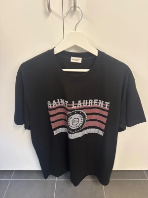 Svart Saint Laurent t-shirt med tryck - Cool svart t-shirt från Saint Laurent med ett stort tryck framtill i vitt och rött. Klassisk rund halsringning och korta ärmar. Perfekt för dig som vill ha en trendig och stilren look med exklusiv känsla.