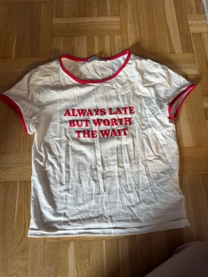 Vit t-shirt med röd text och kant - Vit t-shirt med röd text 'ALWAYS LATE BUT WORTH THE WAIT' på bröstet. T-shirten har röda kanter vid halsringning och ärmslut, samt en klassisk passform och korta ärmar. Perfekt för en avslappnad och trendig stil.