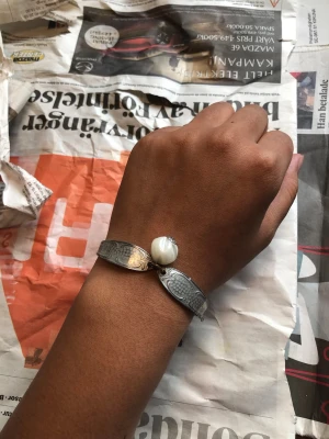 Silverfärgat armband med vit pärla - Snyggt armband i silverfärg med graverade detaljer och en stor vit (plast) pärla i  mitten. Armbandet har en unik design med två böjda metallskaft av bestick som möts vid pärlan. Perfekt accessoar för att lyfta din stil.