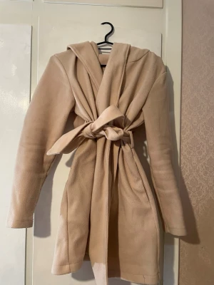 Beige kappa med huva och knytskärp - Stilren beige kappa med stor huva och knytskärp i midjan. Kappan har raka ärmar och två sidofickor. Tillverkad i mjukt, filtliknande material som ger en avslappnad men ändå trendig look.