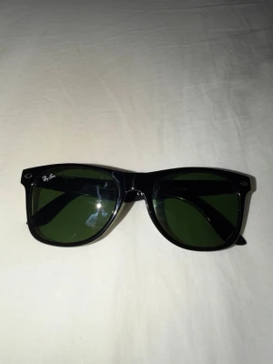 Svarta Ray-Ban Wayfarer solglasögon - Klassiska svarta Wayfarer solglasögon från Ray-Ban med gröna glas och ikonisk logga på bågen. Snygg och tidlös design med robust plastbåge och silverdetaljer vid gångjärnen. Perfekta för att höja din stil i solen.