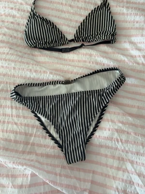 Svartvit randig bikini - Snygg bikini med svartvita vertikala ränder. Överdelen har trekantskupor och justerbara band med spänne i ryggen. Underdelen är klassisk med hög benskärning och samma randiga mönster. Perfekt för stranden eller poolen. 