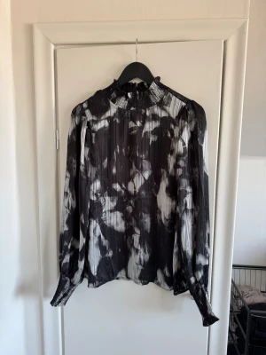 Svartvit plisserad blus med hög krage - Snygg svartvit blus med abstrakt mönster och plisserat tyg. Blusen har hög smockad krage och långa ärmar med smock vid ärmsluten. Materialet ser ut att vara lätt och något transparent, vilket ger en luftig känsla.