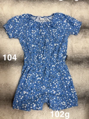 Blå blommig jumpsuit med kort ärm - Supersöt blå jumpsuit med vita småblommor och korta ärmar. Resår i midjan för skön passform och avslappnad look. Perfekt för varma dagar och enkel att styla med sneakers eller sandaler.🌸 Använd en gång. Storlek 104 