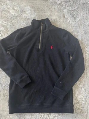Svart stickad tröja med dragkedja - Svart stickad tröja från Polo Ralph Lauren med halv dragkedja och röd broderad logga på bröstet. Tröjan har hög krage och långa ärmar, perfekt för lager på lager. Klassisk och stilren design som funkar till det mesta.