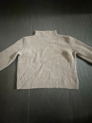 Beige ribbstickad tröja från Wera - Snygg beige ribbstickad tröja från Wera med hög krage och långa ärmar. Tröjan har en rak passform och är tillverkad i ett mjukt, tjockare material som passar perfekt för kyligare dagar. Enkel och stilren design som är lätt att matcha.