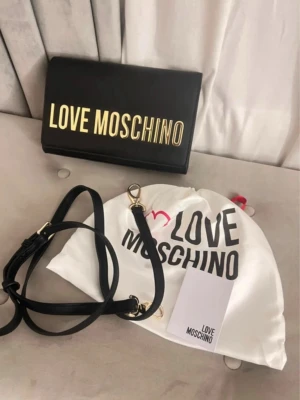 Moschino - Snygg och elegant axelväska från Love Moschino i svart med guldlogga framtill. Perfekt både till vardag och fest. Väskan är i mycket fint nytt skick och knappt använd:  Medföljer: Guldkedja axelrem  Extra svart axelrem  Original dustbag Mått:  Höjd: 14 cm  Längd: 22 cm  Djup: 5,5 cm  Axelrem: ca 55 cm 