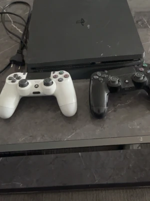 PlayStation 4 Slim med två kontroller - Säljer en svart PlayStation 4 Slim från Sony tillsammans med två DualShock 4-kontroller, en vit och en svart. Konsolen har en stilren, rektangulär design och är tillverkad i plast. Perfekt för gaming med vänner och familj.