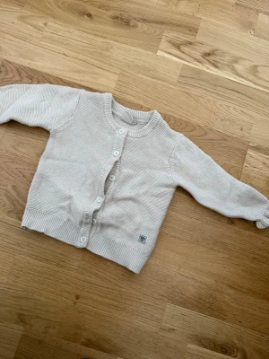 Beige stickad kofta barn - Söt beige stickad kofta för barn i storlek 92. Koftan har rund hals, långa ärmar och stängs med vita knappar framtill. Ribbstickade muddar vid ärmslut och nederkant ger en klassisk look. Perfekt för lager på lager under kyliga dagar.