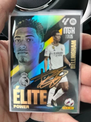 Panini Elite Power Bellingham kort - Samlarbild från Panini med fotbollsspelaren Bellingham i Real Madrid. Kortet har holografisk effekt, guldtext och spelarens autograftryck. Framsidan visar Bellingham i matchställ och baksidan har statistik och info på spanska. Perfekt för samlare!