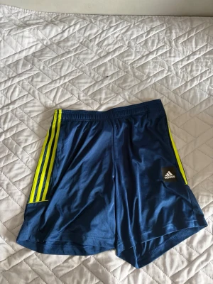 Blå Adidas shorts med gula ränder - Snygga blå träningsshorts från Adidas med klassiska gula ränder längs sidorna och Adidas-logga på ena benet. Shortsen har elastisk midja och är gjorda i ett lätt och luftigt syntetmaterial, perfekta för sport eller gym.