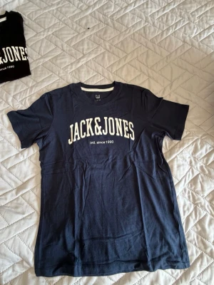Mörkblå Jack & Jones t-shirt - Snygg mörkblå t-shirt från Jack & Jones med vit logga tryckt på bröstet. Klassisk rund halsringning och korta ärmar. Perfekt för en avslappnad och stilren look.