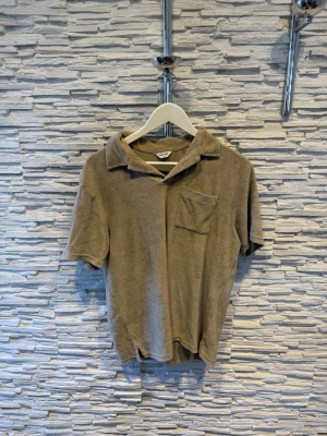Beige frottétopp med krage - Beige topp i mjuk frotté med klassisk krage och korta ärmar. Toppen har en bröstficka och avslappnad passform, perfekt för en chill och trendig stil.