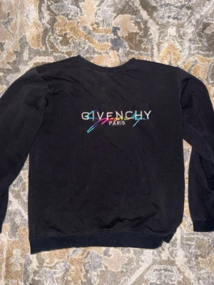 Svart Givenchy sweatshirt - Säljer en svart sweatshirt från Givenchy med färgglad broderad logga och texten 'Paris' på bröstet. Tröjan har rund halsringning och ribbade muddar vid ärmslut och nederkant. Perfekt för dig som gillar streetwear och exklusiva märken.