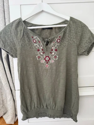 Olivgrön blus med broderi och knyt - Olivgrön blus med korta ärmar, broderade blommor i vitt och rött framtill samt dekorativ knytning vid halsen. Ärmarna har hålmönster och blusen har resår i nederkant för en avslappnad passform.