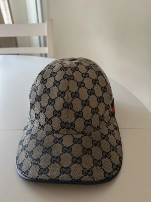 Zara - Snygg beige keps med klassiskt GG-monogram över hela kepsen. Kepsen har böjd skärm och en detalj med röd och marinblå rand på sidan. Tillverkad i canvasmaterial med exklusiv känsla och modern passform.