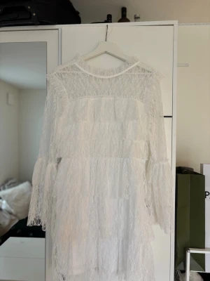 Vit spetsklänning med volanger - Otroligt fin vit klänning i spets med långa ärmar och volangdetaljer. Klänningen har en lätt transparent look och rund halsringning. Perfekt för dig som gillar en feminin och bohemisk stil. Klänningen är en  studentklänning som aldrig kom till användning, den är aldrig använd förutom vid prov.