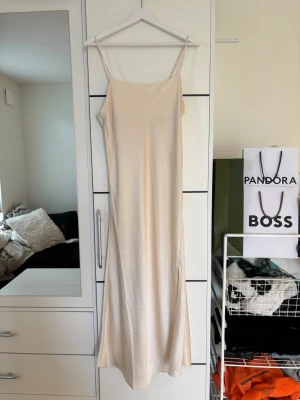 Lång champagnefärgad slipklänning - Säljer en elegant lång slipklänning i champagnefärg med tunna axelband och slits i sidan. Klänningen är i satin och ger en mjuk, glansig yta som ger en lyxig känsla och faller snyggt längs kroppen. Perfekt för dig som gillar minimalistisk och stilren design. Klänningen är köpt på Kappahl i storlek S!