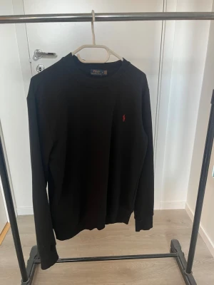 Svart tröja från Polo Ralph Lauren - Klassisk svart långärmad tröja från Polo Ralph Lauren med rund halsringning och den ikoniska röda logotypen broderad på bröstet. Tröjan har ribbade muddar vid ärmslut och nederkant. Perfekt för en stilren och enkel look.