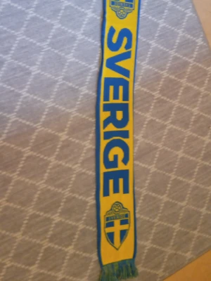 Gul och blå Sverige-halsduk - Supporterscarf i gult och blått med texten 'Sverige' och svenska landslagets emblem på båda sidor. Halsduken har blå fransar i ändarna och är perfekt för att visa ditt stöd på matcher eller event. Tillverkad i mjukt material.