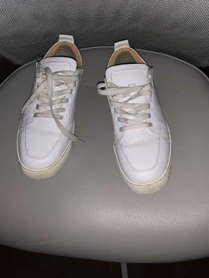 Vita sneakers med röd sula - Stilrena vita sneakers med lågt skaft och klassisk snörning. Skorna har en unik röd sula med mönster och är tillverkade i slätt läder. Perfekta för dig som gillar minimalistisk design med en twist. Jag har tyvär tappat bort kartongen och alla kvitto bevis och sånt men har en påse där de står Louboutin som man får när man köper