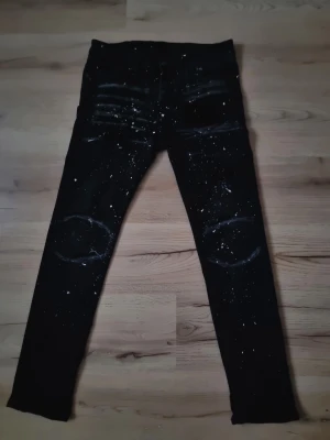 Svarta skinny jeans med färgstänk - Säljer ett par svarta skinny jeans med coola färgstänk och slitningar över knäna. Jeansen har en tight passform och detaljerade sömmar vid låren och knäna som ger en edgy look. Perfekta för dig som vill sticka ut med din stil.