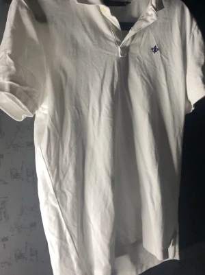 MORRIS PIKÉTRÖJA  - Klassisk vit pikétröja med korta ärmar och krage. Tröjan har en liten broderad detalj på bröstet och knäppning vid halsen. Perfekt till jeans eller shorts för en clean och enkel stil.