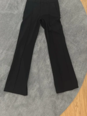 Svarta utsvängda kostymbyxor - Säljer ett par svarta kostymbyxor med utsvängda ben och pressveck. Byxorna har hög midja och klassisk passform, perfekta för en stilren look. Materialet ser ut att vara en mjuk och följsam mix, troligtvis polyester eller viskos.