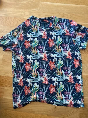 Blommig t-shirt från Jack & Jones - Säljer en färgstark blommig t-shirt från Jack & Jones Premium. T-shirten har korta ärmar och ett mönster med blommor i rött, lila, vitt och grönt mot en mörkblå bakgrund. Perfekt för dig som vill sticka ut med en tropisk vibe.