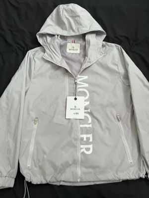 Ljusgrå vindjacka från Moncler - Snygg ljusgrå vindjacka från Moncler med huva och dragkedja framtill. Jackan har två fickor med dragkedja, stor Moncler-logga tryckt vertikalt på framsidan och ett Moncler-märke på ärmen. Tillverkad i lätt, vindtätt material som passar perfekt till streetwear.