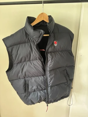 Vändbar dunväst från Fjällräven - Säljer en svart dunväst från Fjällräven med hög krage och dragkedja. Västen har flera praktiska fickor framtill och den klassiska Fjällräven-loggan på bröstet. Perfekt för lager-på-lager och outdoor-looks. Men även en dunfunktion.