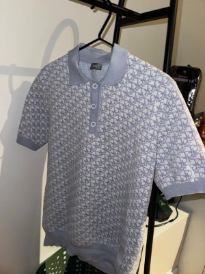 DR Piké T-shirt - Snygg ljusblå piké t-shirt med korta ärmar och ett diskret mönster över hela tröjan. Tröjan har krage, knappar framtill och ribbade muddar vid ärmslut och nederkant. Perfekt för en stilren och avslappnad look.