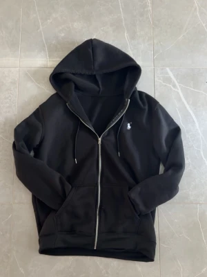 Svart zip hoodie med logga - Säljer en svart hoodie med dragkedja och huva. Tröjan har två fickor framtill och en vit broderad logga på bröstet. Mjuk insida och justerbara snören i huvan. Perfekt för en avslappnad och stilren look.