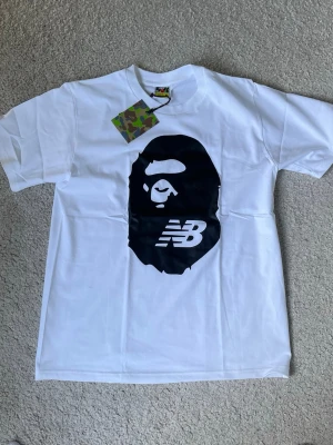 Vit BAPE x New Balance t-shirt - Säljer en vit t-shirt från BAPE x New Balance med den ikoniska BAPE-apan och NB-loggan tryckt i svart på bröstet. Klassisk passform och rund hals. T-shirten har en liten BAPE-tag på ärmen och är tillverkad i mjuk bomull.