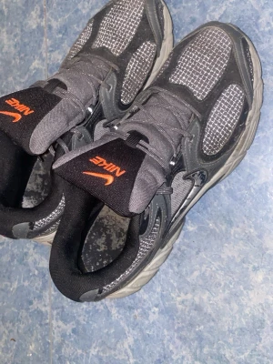 Nike svarta och grå sneakers - Snygga sneakers från Nike i svart och grått med meshpaneler och detaljer i mockaimitation. Skorna har en sportig look med orange Nike-logga på plösen och robust sula. Perfekta för dig som gillar streetwear och vill ha bekväma skor till vardags.