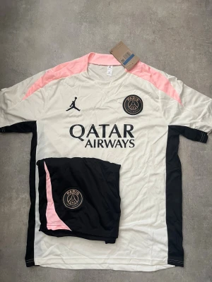 PSG matchställ i vitt och svart - Säljer ett PSG matchställ med tröja och shorts. Tröjan är vit med svarta och rosa detaljer, PSG-logga och Jordan-logga på bröstet samt Qatar Airways-tryck. Shortsen är svarta med PSG-logga och rosa detalj. Perfekt för fotbollsträning eller match.