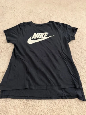 Svart Nike t-shirt - Snygg svart t-shirt från Nike med stor vit logga på bröstet. Klassisk rund halsringning och korta ärmar, och T-shirten är lite kortare framtill (bild 1). Tillverkad i mjuk bomull som känns skön mot huden. (Kolla tredje bilden för att se korrekt storlek)💕