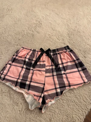 Pyjamasshorts  - Snygga pyjamasshorts i rosa med svart rutmönster och en söt svart rosett framtill. Tillverkade i mjukt material med 95% polyester och 5% elastan för extra komfort. Perfekta för mysiga kvällar hemma. Helt oanvända 
