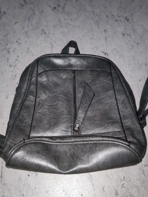 Grå ryggsäck i konstläder 29x29 - Snygg grå ryggsäck i konstläder med en framficka med dragkedja och justerbara axelband. Väskan har en enkel, stilren design och passar perfekt för dig som vill ha en praktisk och trendig accessoar. Röd insida syns på sidan.