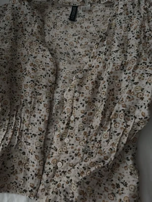 Blommig blus från H&M Divided - Söt blommig blus från H&M Divided med små beige och bruna blommor på ljus botten. Blusen har knappar framtill, v-ringning och långa ärmar med lätt puff och rynk vid axlarna. 