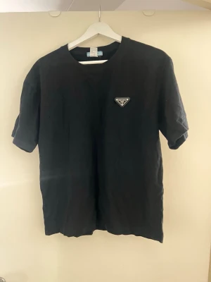 Svart t-shirt från Prada i bomull - Svart t-shirt från Prada med klassisk rund hals och korta ärmar. Diskret Prada-logga på bröstet i form av en liten patch. Tillverkad i mjuk bomull för en skön och stilren look.
