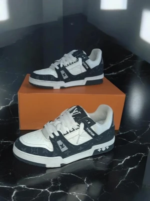 Louis Vuitton trainers svart och vitt sneakers - Snygga Louis Vuitton sneakers i vitt och mörkgrått med klassiskt monogrammönster. Skorna har lågt skaft, vita snören och detaljerade LV-loggor på sidan och tungan. Tillverkade i läder och textil för en lyxig känsla. Perfekta för dig som gillar exklusiv streetstyle.