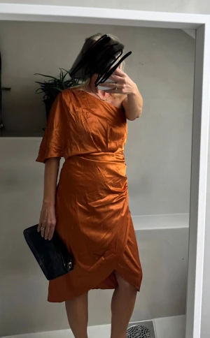 Kopparfärgad omlottklänning i satin - Snygg kopparfärgad omlottklänning i glansigt satinmaterial. Klänningen har en asymmetrisk v-ringning, kort ärm på ena sidan och omlottdetalj med drapering i midjan. Perfekt för dig som vill sticka ut med en unik look.