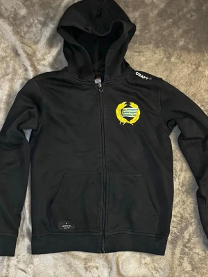 Svart Hammarby hoodie från Craft - Svart hoodie med dragkedja från Craft, prydd med Hammarby IF:s klubbmärke i grönt, vitt och gult på bröstet. Huva och två fickor framtill. Perfekt för dig som vill representera laget med stil. Vid frågor hör av dig och priset kan alltid diskuteras 