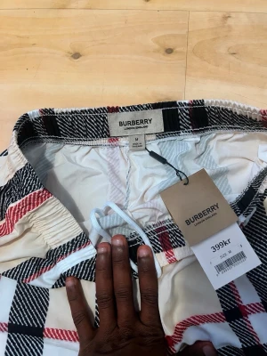 Rutig Burberry shorts i bomull - Säljer ett par klassiska rutiga shorts från Burberry i beige, svart, vit och röd. Shortsen har elastisk midja och är tillverkade i mjuk bomull. Perfekta för sommaren och har den ikoniska Burberry-looken.